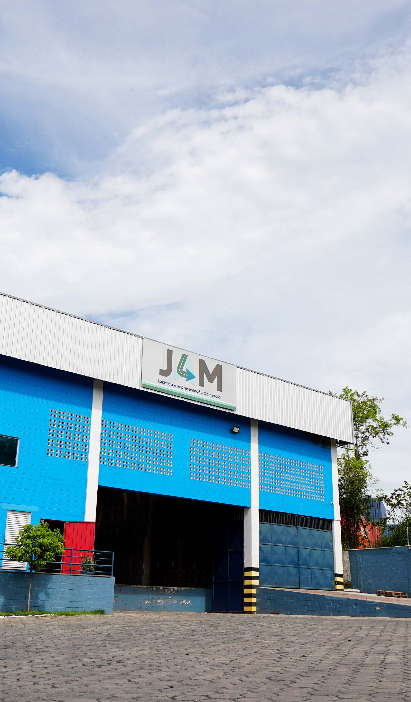 Fachada da unidade da JAM Logística pintada em azul, com logotipo e âncora da marca, caixa-d'água e pátio pavimentado sob céu de nuvens